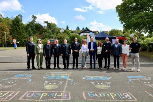Gruppe von Ehrengästen und Feuerwehrangehörigen auf einem Schulhof, im Vordergrund mit Straßenkreide gemalte Werte-Begriffe wie „Frieden“ und „Bildung“