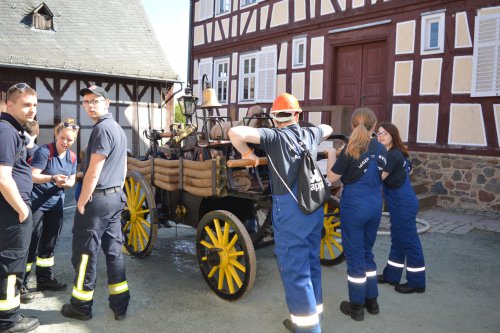 Feuerwehrleute und Jugendliche betrachten eine historische Handdruckspritze mit gelben Holzrädern vor einem Fachwerkhaus