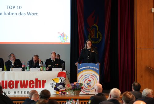 Frau im Dienstanzug spricht am Rednerpult, daneben das sitzende Präsidium, an der Wand die Folie „TOP 10 – Gäste haben das Wort“