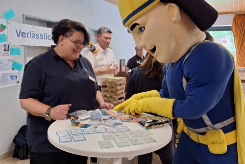 Maskottchen im Superhelden-Kostüm spielt mit einer Betreuerin am Stehtisch ein Wertekarten-Spiel, daneben ein Jenga-Turm