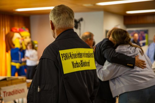 Mitglied des PSNV-Kriseninterventionsdienstes Marburg-Biedenkopf von hinten, im Hintergrund umarmen sich zwei Personen