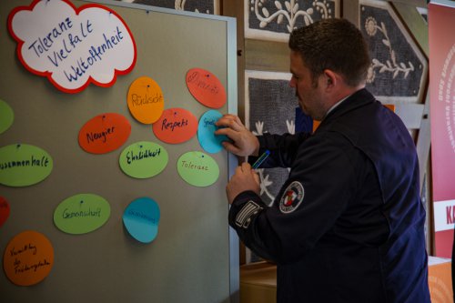 Feuerwehrmann heftet an einer Stellwand zum Thema „Toleranz, Vielfalt, Weltoffenheit“ eine beschriftete Karte an, daneben Begriffe wie Respekt, Neugier und Ehrlichkeit
