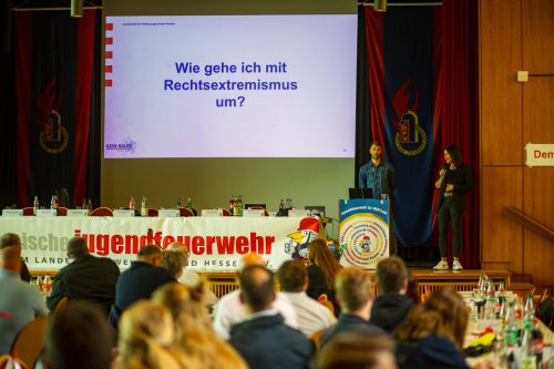 Zwei Vortragende auf der Bühne vor den Delegierten, an der Wand die Folie „Wie gehe ich mit Rechtsextremismus um?“