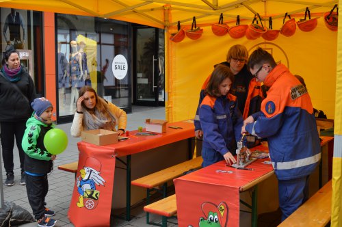 Kinder probieren an einem Mitmachstand mit aufgehängten Helmen unter Anleitung der Jugendfeuerwehr etwas aus, daneben ein Junge mit grünem Luftballon