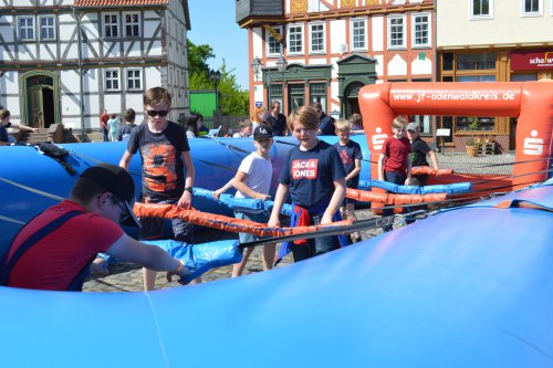 Jugendliche spielen Menschenkicker auf einem aufblasbaren blauen Spielfeld vor Fachwerkhäusern