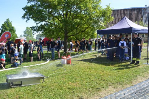 Kinder und Jugendliche verfolgen unter einem Baum eine Vorführung mit Feuerwehrschlauch und Spritze auf der Wiese