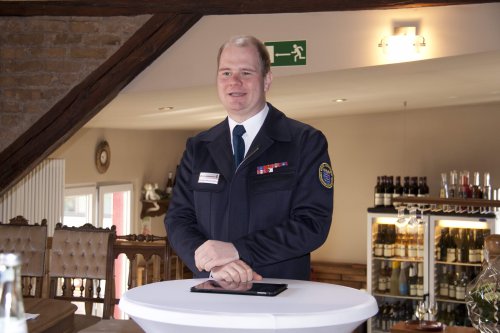 Mann in blauer Feuerwehruniform steht lächelnd an einem Stehtisch und moderiert die Verabschiedung