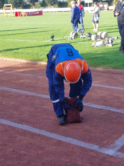 Wettkämpfer mit Startnummer 9 und orangefarbenem Helm legt auf der Aschenbahn eine Schlauchtasche ab