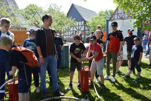 Kinder bedienen gemeinsam mit Betreuern rote Kübelspritzen auf einer Wiese vor Fachwerkhäusern
