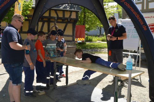 Jugendlicher balanciert unter einem Pavillon auf einer Holzplanke, umringt von Kindern und Betreuern