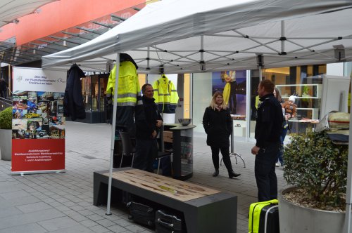 Mehrere Personen in gelber Warnkleidung an einem Infostand mit Roll-up zur Feuerwehr-Ausbildung im Einkaufszentrum