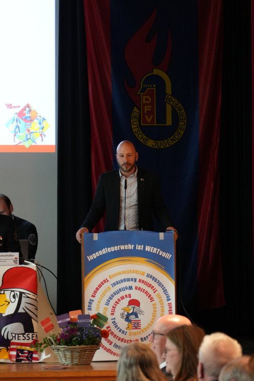 Redner am Pult mit dem Banner „Jugendfeuerwehr ist WERTvoll!“ vor einer Fahne der Deutschen Jugendfeuerwehr