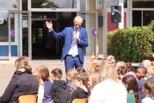 Bildungsminister Armin Schwarz spricht mit Mikrofon zu einer Gruppe sitzender Grundschulkinder vor dem Schuleingang