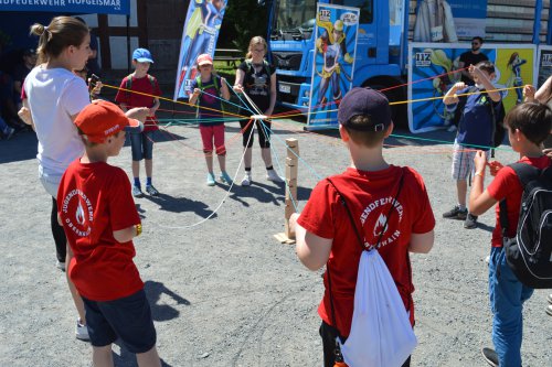 Kinder in roten Jugendfeuerwehr-Shirts spannen im Kreis ein Netz aus bunten Schnüren bei einem Teamspiel