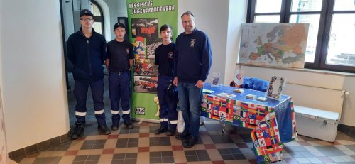 Drei Jugendliche und ein Betreuer der Hessischen Jugendfeuerwehr am Messestand mit Europakarte und einem mit Länderflaggen geschmückten Tisch