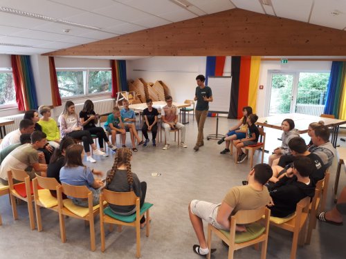 Jugendliche sitzen im Stuhlkreis in einem Gruppenraum, in der Mitte spricht ein Betreuer; an der Wand Vorhänge in Regenbogen- und Deutschland-Farben