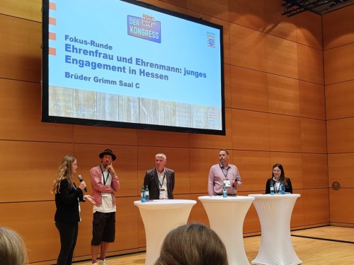 Fokus-Runde „Ehrenfrau und Ehrenmann: junges Engagement in Hessen“ auf der Bühne des Ehrenamtskongresses; fünf Diskussionsteilnehmende an Stehtischen vor der Leinwand