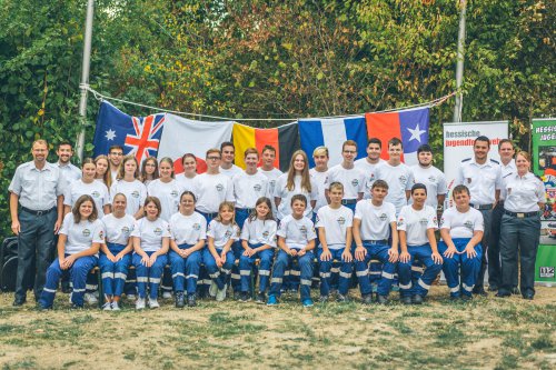 Gruppenfoto der Teilnehmenden der internationalen Jugendbegegnung vor einer Reihe gehisster Länderflaggen, dahinter Banner der Hessischen Jugendfeuerwehr