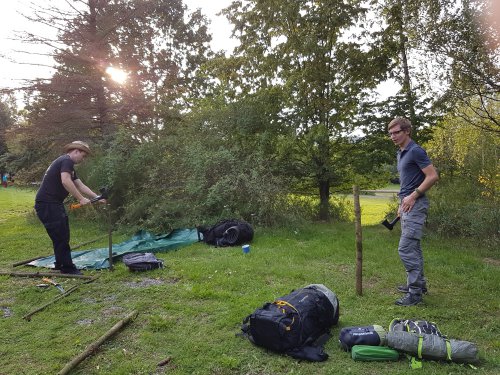 Teilnehmer bauen auf einer Wiese ein Tarp als Unterschlupf auf, daneben liegen Rucksäcke und Schlafsäcke