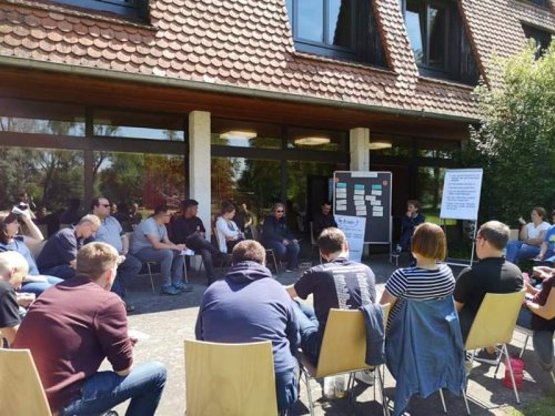 Workshop-Teilnehmende sitzen im Stuhlkreis im Freien vor einem Gebäude, daneben Stellwände mit Moderationskarten