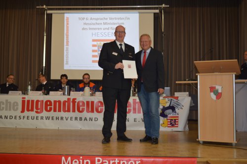 Ehrung auf der Bühne: zwei Männer mit Urkunde, einer in Feuerwehruniform