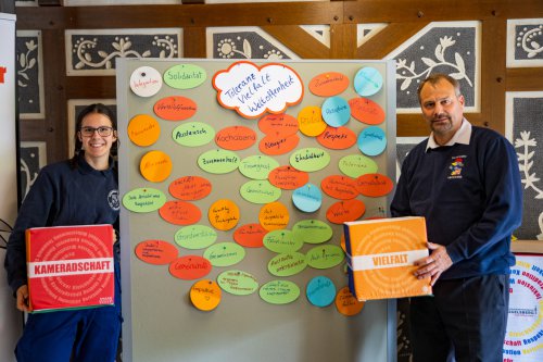 Zwei Personen präsentieren neben einer voll beklebten Stellwand zum Thema „Toleranz, Vielfalt, Weltoffenheit“ und halten Kartons mit den Aufschriften „Kameradschaft“ und „Vielfalt“