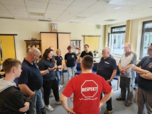 Teilnehmende stehen im Stuhlkreis bei einem Workshop, in der Mitte eine Person im roten T-Shirt mit Aufdruck „Respekt"