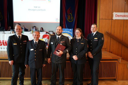 Gruppenbild von fünf Geehrten in Dienstuniform vor der Bühne, eine Person hält eine Urkundenmappe, an der Wand die Folie „TOP 13 – Ehrungen und Dank“