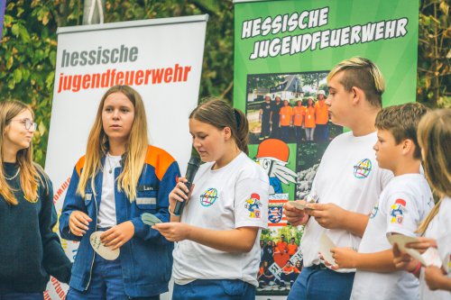 Jugendliche präsentieren beim Abschlussabend ihre Ergebnisse, ein Mädchen spricht ins Mikrofon, im Hintergrund Banner der Hessischen Jugendfeuerwehr