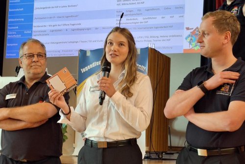 Junge Frau spricht in ein Mikrofon und hält einen Bausatz hoch, flankiert von zwei zuhörenden Männern in Feuerwehrkleidung