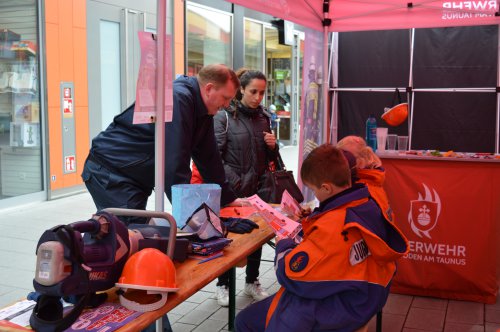 Ein Junge in Jugendfeuerwehr-Kleidung bastelt an einem Stand, daneben ein Erwachsener und Besucher, auf dem Tisch eine Stichsäge und ein Helm