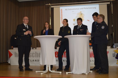 Podiumsrunde an Stehtischen auf der Bühne, im Hintergrund die Präsentation „AG Extremismus und Polarisierung“