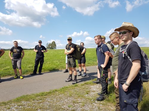 Gruppe des Outdoor-Lehrgangs steht mit Rucksäcken auf einem Feldweg und lauscht den Erklärungen