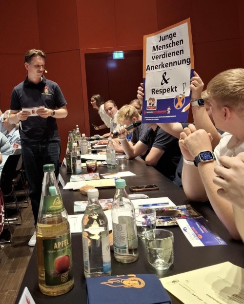 Teilnehmende halten an einem Veranstaltungstisch ein selbst gestaltetes Wahlplakat mit der Aufschrift „Junge Menschen verdienen Anerkennung & Respekt“ hoch