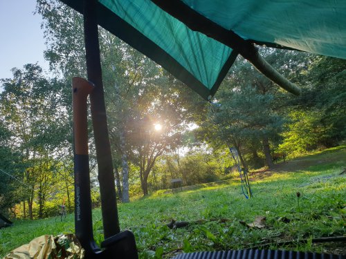 Blick aus dem Inneren eines aufgespannten Tarps in die Abendsonne, im Vordergrund eine Axt