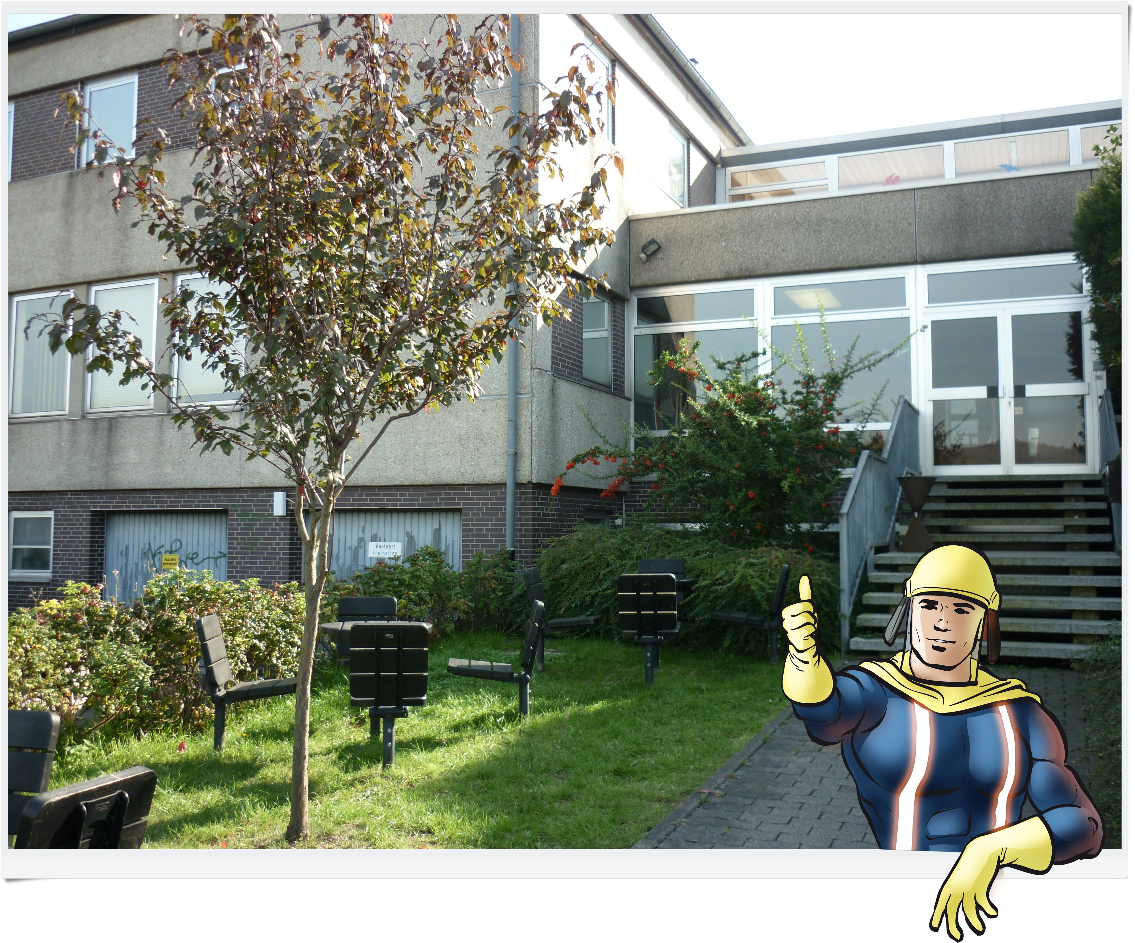 Außenansicht des Jugendfeuerwehr-Ausbildungszentrums mit Grünfläche, davor ein eingefügtes Comic-Maskottchen mit Daumen hoch