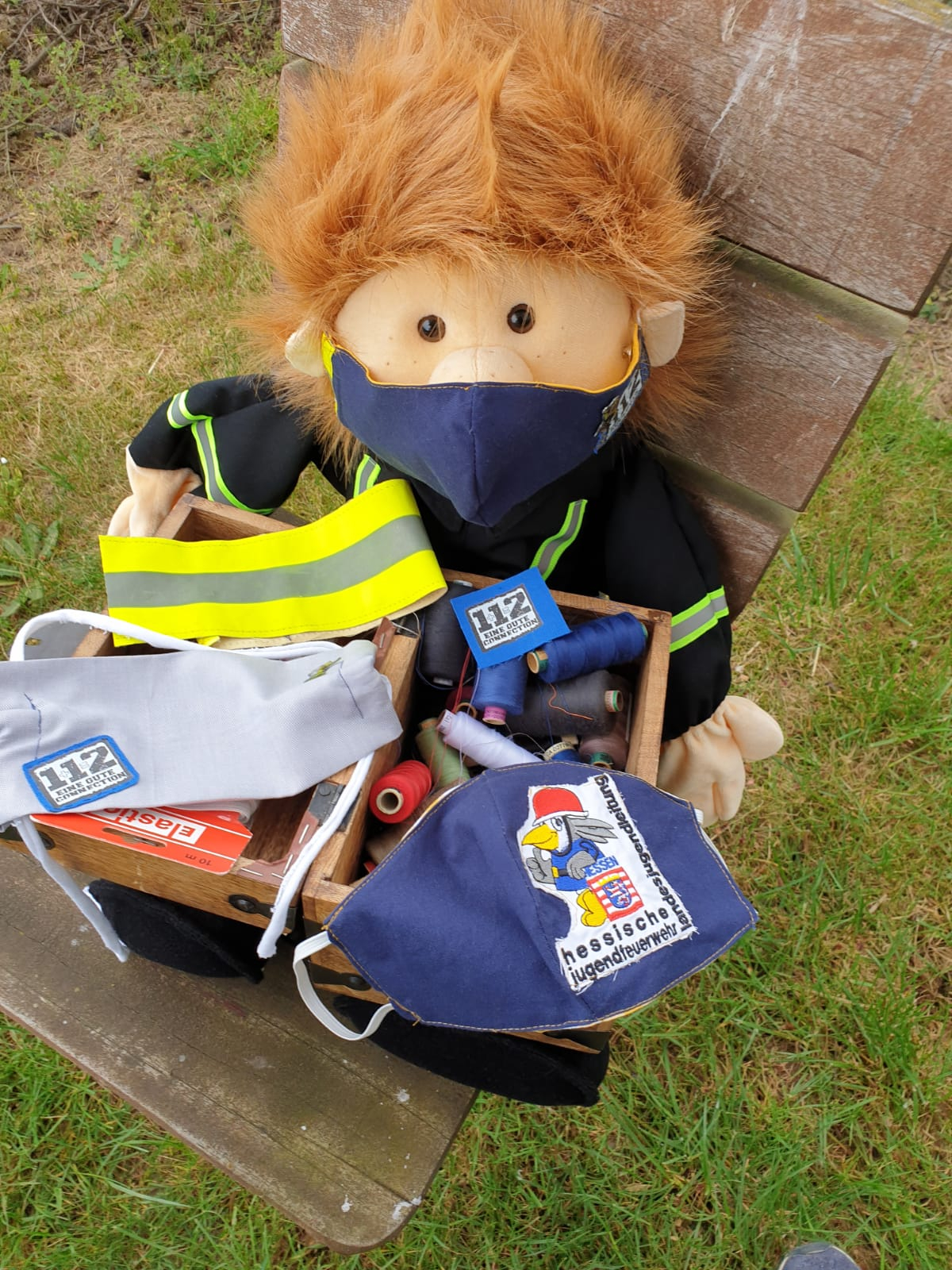 Plüsch-Maskottchen mit Feuerwehrjacke und Mund-Nasen-Bedeckung neben einer Kiste mit selbstgenähten Behelfsmasken