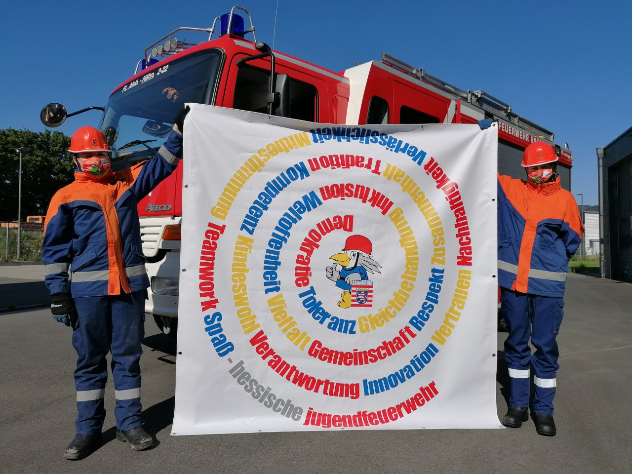 Zwei Jugendfeuerwehrmitglieder in Uniform halten vor einem Feuerwehrfahrzeug die Wertespirale-Fahne der Hessischen Jugendfeuerwehr mit Begriffen wie Demokratie, Toleranz und Respekt