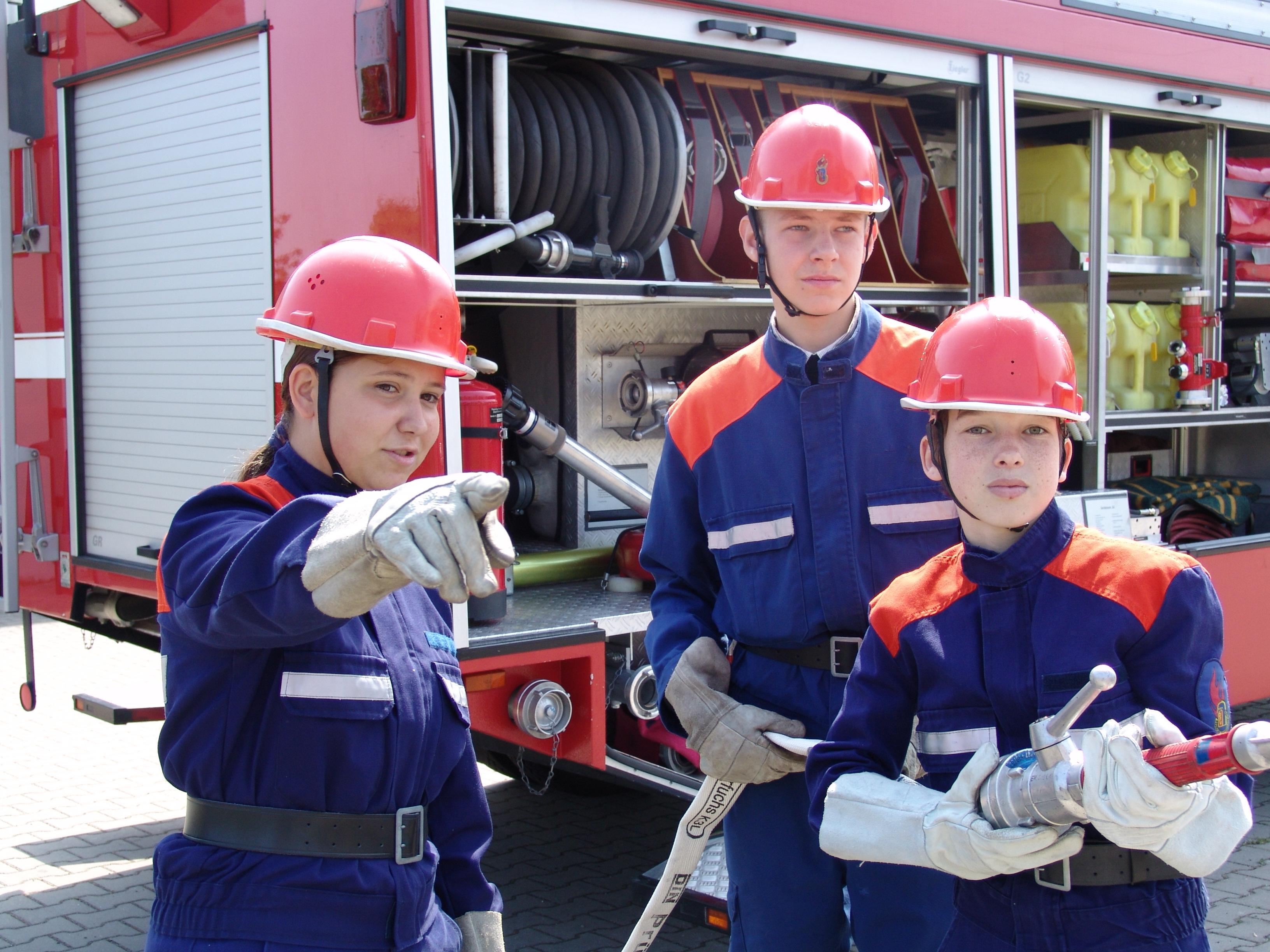 Drei Jugendfeuerwehrmitglieder in blauer Uniform mit roten Helmen stehen mit Strahlrohr und Schlauch vor einem geöffneten Feuerwehrfahrzeug