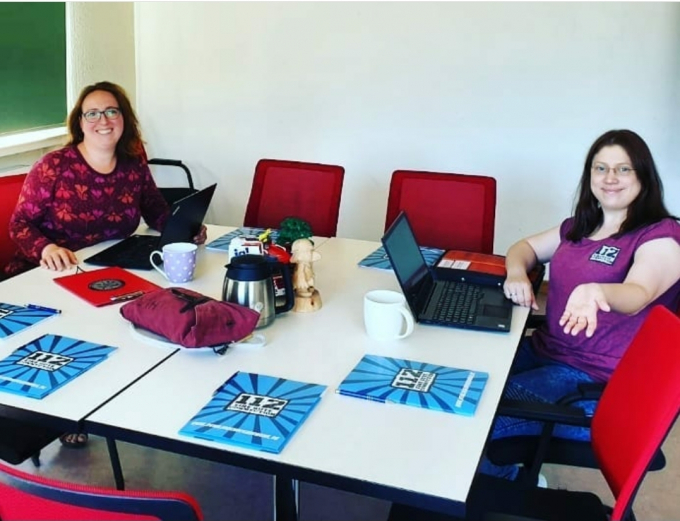 Zwei lächelnde Frauen sitzen mit Laptops an einem Besprechungstisch mit Schulungsunterlagen und Tassen