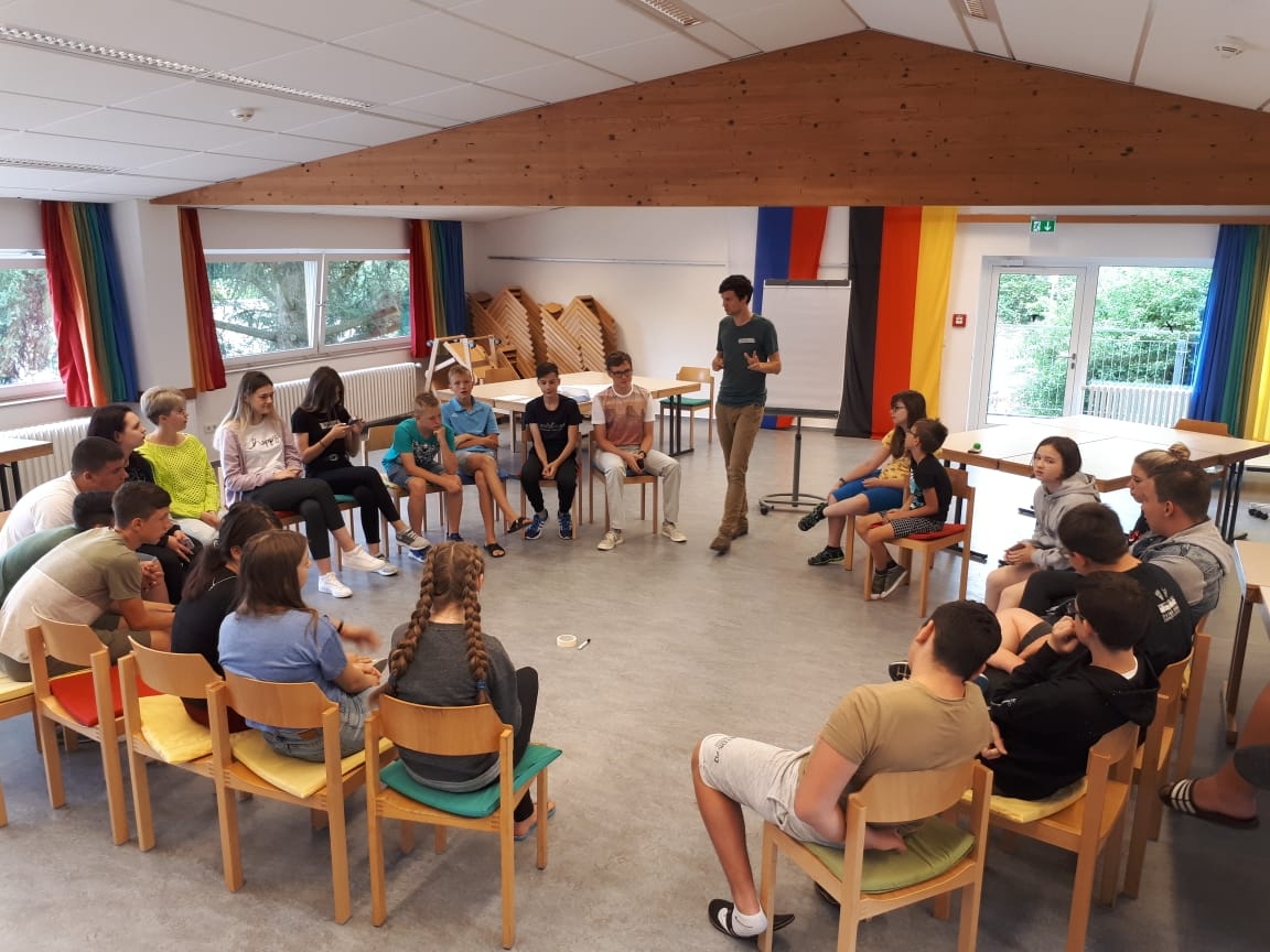 Jugendliche sitzen im Stuhlkreis in einem Seminarraum mit Holzdecke, eine Person steht und spricht zur Gruppe