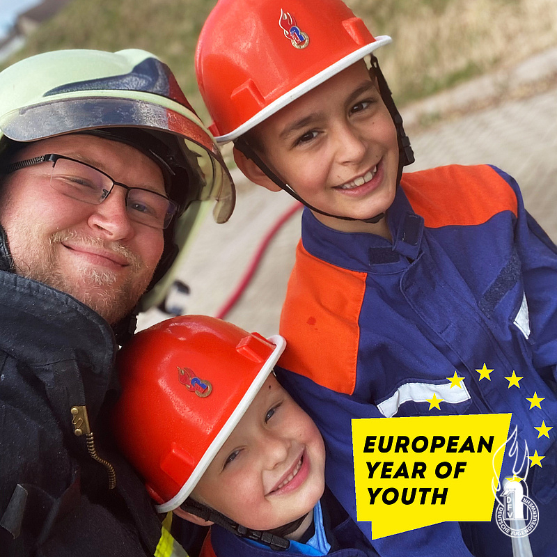 Feuerwehrmann mit zwei lachenden Kindern in Jugendfeuerwehr-Helmen, daneben der Schriftzug „European Year of Youth"