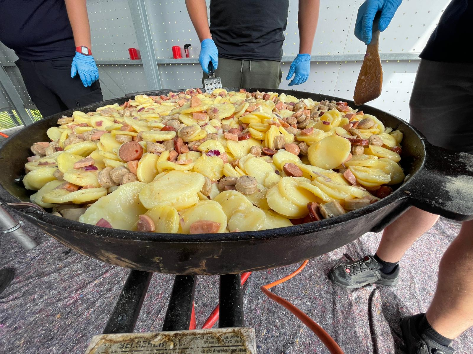 Riesige Gusseisenpfanne mit Bratkartoffeln und Wurststücken, um die mehrere Personen mit Einweghandschuhen beim Kochen im Zeltlager stehen