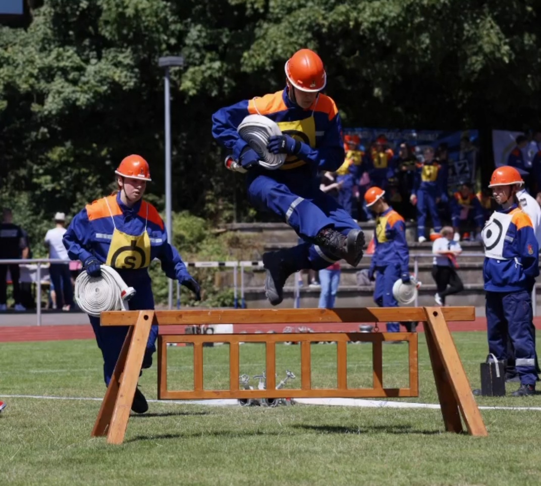 Jugendfeuerwehrmitglied in blauer Uniform und orangem Helm springt beim Wettbewerb mit einem Schlauchpaket über eine Holzhürde, weitere Teilnehmende im Hintergrund