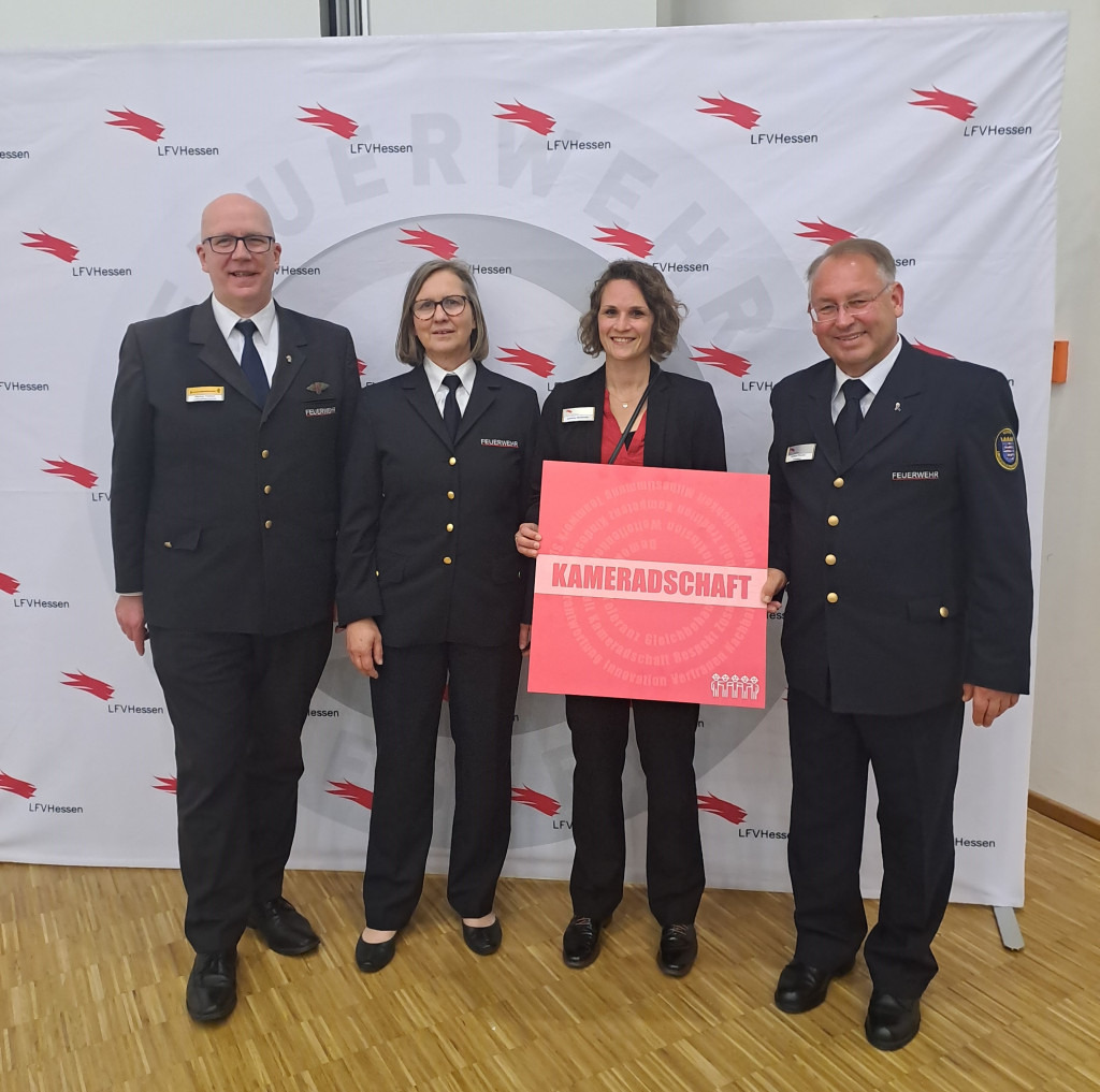 Vier Personen in Feuerwehr-Uniform stehen vor einer LFV-Hessen-Fotowand; eine hält ein rotes Schild mit dem Schriftzug „KAMERADSCHAFT" in den Händen