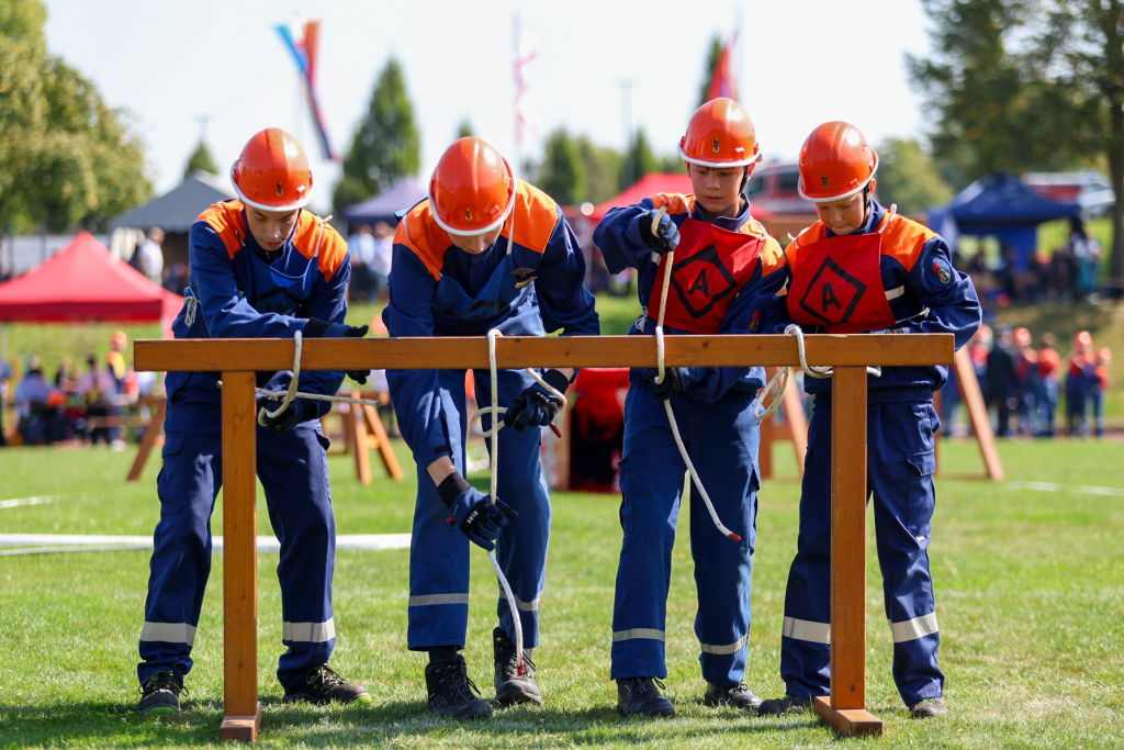 Vier Jugendfeuerwehrmitglieder in blauer Uniform und orangem Helm knoten beim Wettbewerb konzentriert Leinen an einem Holzgestell, im Hintergrund das Wettkampfgelände
