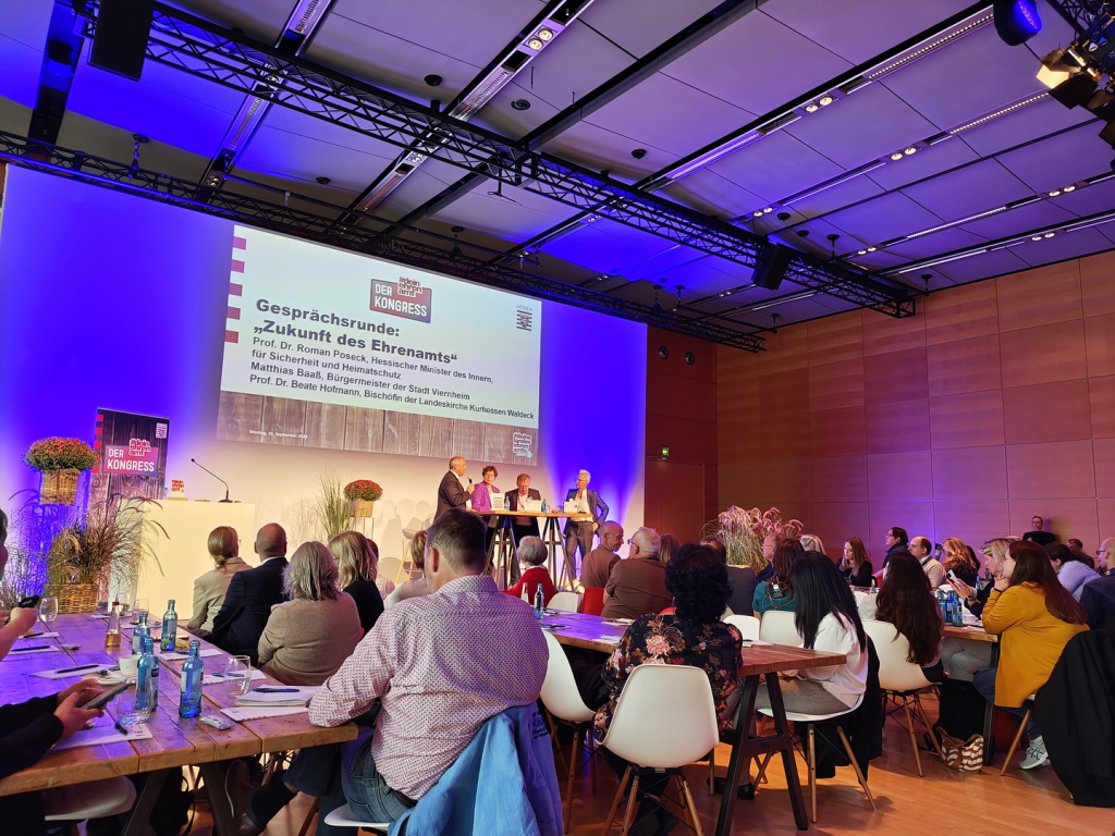 Blau beleuchteter Veranstaltungssaal des Ehrenamtskongresses mit Publikum an Tischen und einer Gesprächsrunde „Zukunft des Ehrenamts" auf der Bühne vor der Leinwand