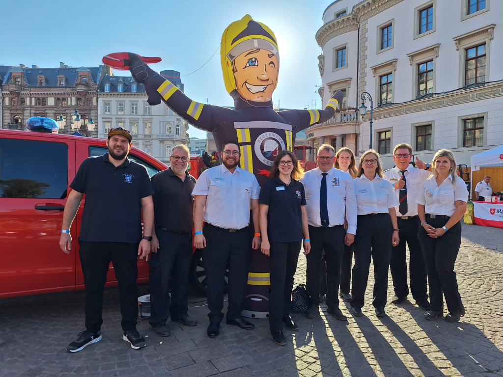 Gruppe von Mitarbeitenden der Hessischen Jugendfeuerwehr posiert vor einem aufblasbaren Feuerwehrmann-Maskottchen und einem roten Einsatzfahrzeug auf einem Platz in der Wiesbadener Innenstadt