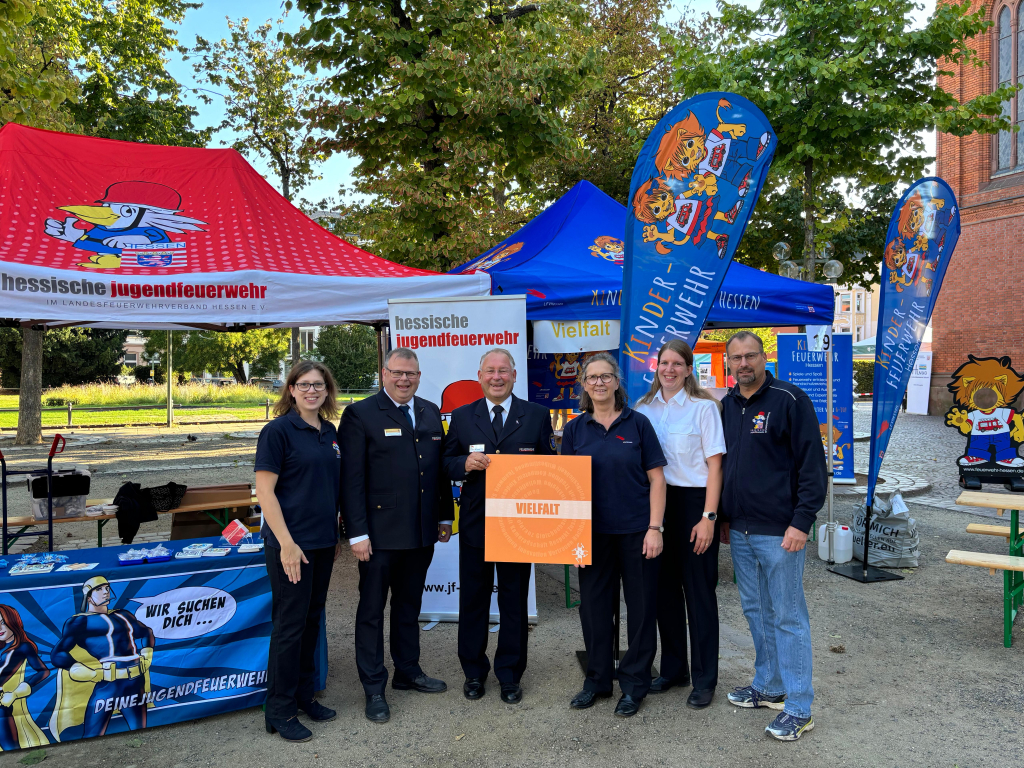 Sechs Personen in Feuerwehrkleidung stehen an einem Infostand der Hessischen Jugendfeuerwehr und der Kinderfeuerwehr und halten einen orangefarbenen Wertedeckel mit dem Schriftzug ‚VIELFALT' in die Kamera.