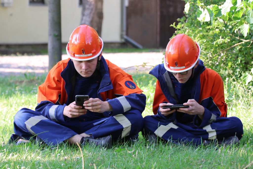 Zwei Jugendliche in Feuerwehruniform mit orangefarbenen Helmen sitzen im Gras und blicken konzentriert auf ihre Smartphones.